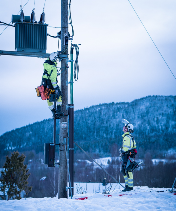 En energimontør klatrer i en kraftmast opp mot en transformator mens en kollega sikrer.